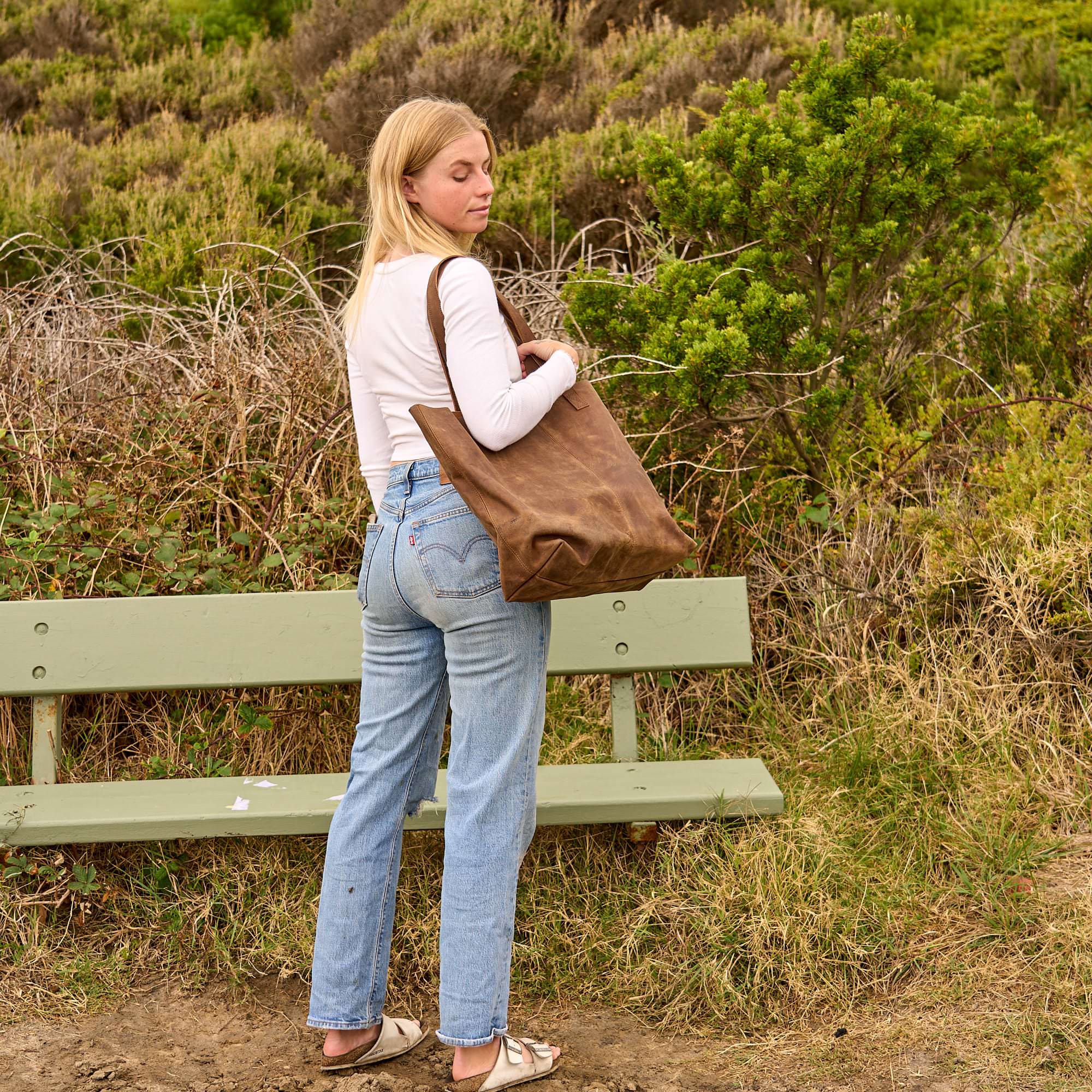 Woman in a white crop top and jeans holding a large vintage brown leather tote bag in front of a white background.

