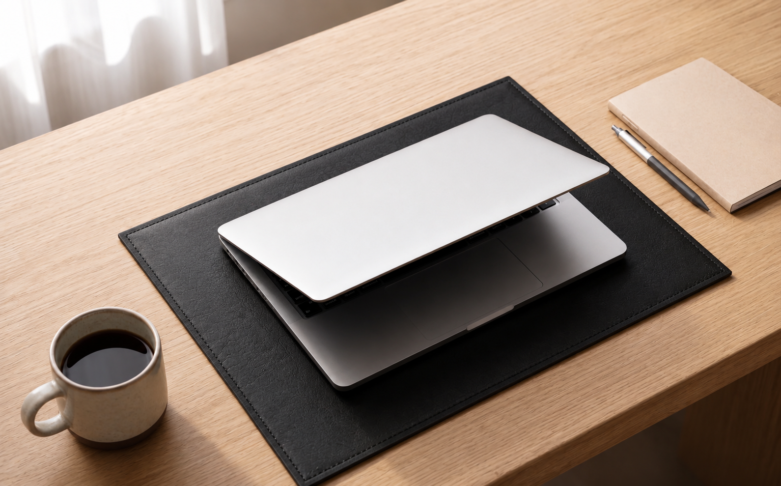 Laptop on a black mat with a cup of coffee and notebook on a wooden desk
